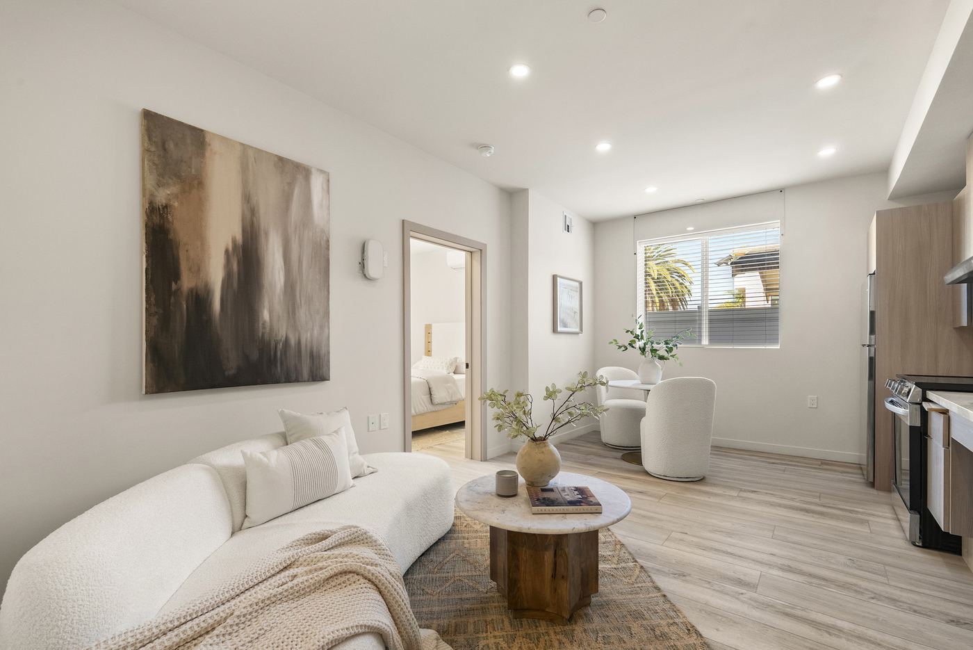 The living area of a ground-floor apartment. A window, an open doorway to the bedroom, and the kitchen along the right wall. Furniture is staged.