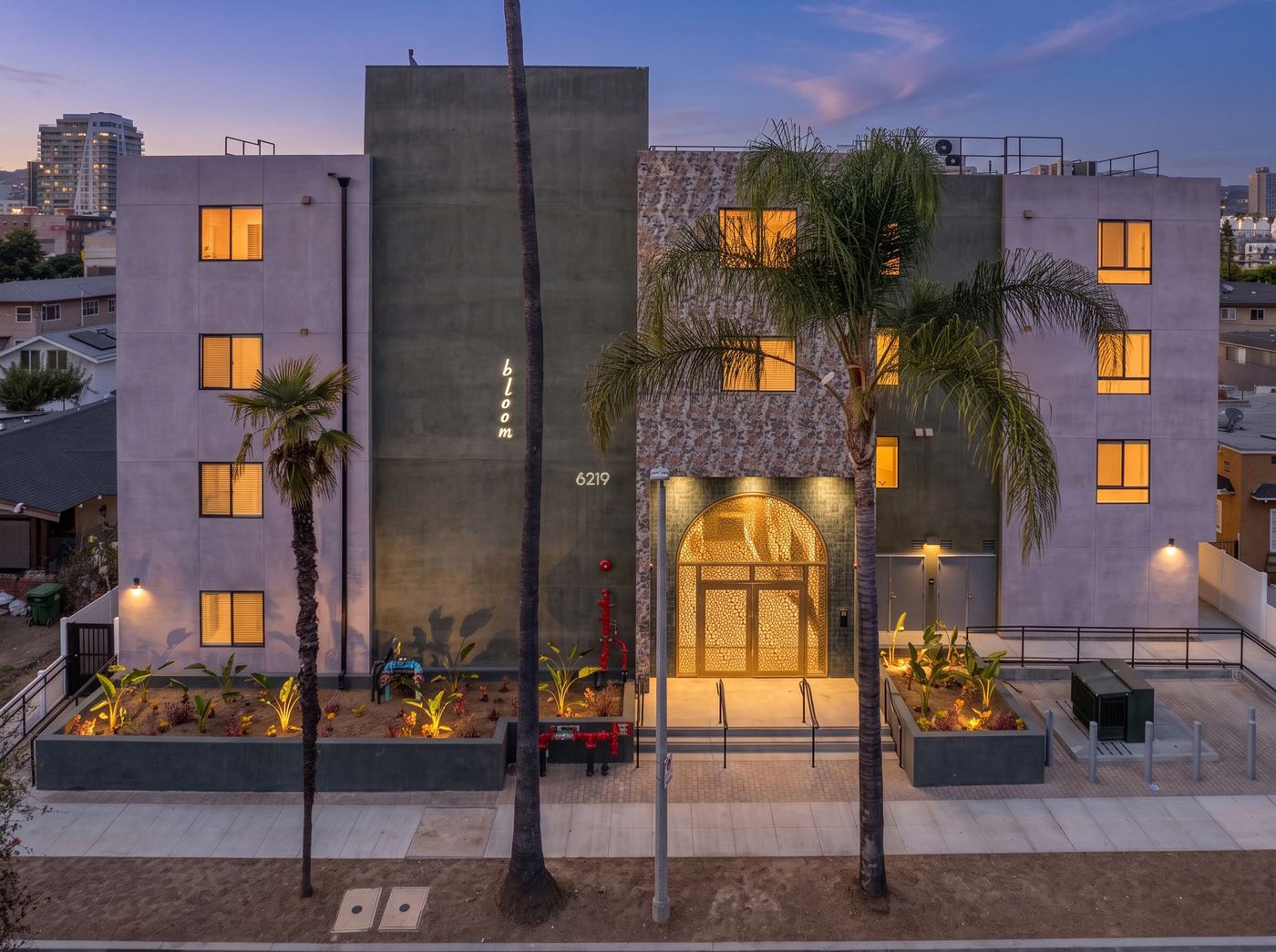 The front of Bloom Apartments at dusk. A four-story building with lit windows, palm trees and planted beds along the sidewalk.